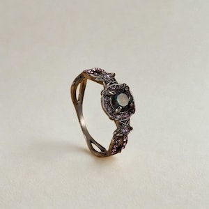 May include: A silver-toned ring with a central dark gemstone, surrounded by smaller pink stones. The band features an intricate, openwork design with additional pink accents. The ring's setting is square, and the overall style is ornate and detailed.