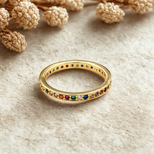 May include: A gold-colored ring with a band of colorful gemstones. The ring features a continuous row of small, round gemstones in various colors, including red, orange, yellow, green, blue, and purple. The ring is set against a neutral background.