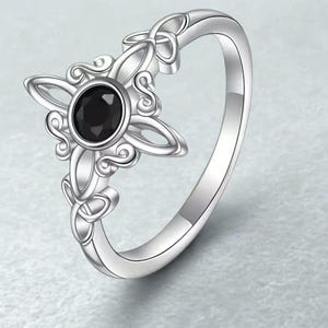 May include: A silver ring with a black gemstone at its centre. The ring showcases a four-petal floral design with detailed knotwork. The band is smooth and polished, reflecting light.
