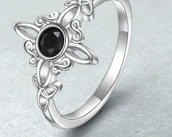 Puede incluir: Un anillo plateado con una piedra negra en el centro. El diseño del anillo incluye un motivo floral de cuatro pétalos con intrincados detalles de nudos. La banda es lisa y pulida, reflejando la luz.