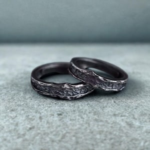 May include: Two textured, dark gray rings. The rings have a hammered metal appearance with a rough, uneven surface. The bands are circular and sit on a light gray surface with a blue-gray background.