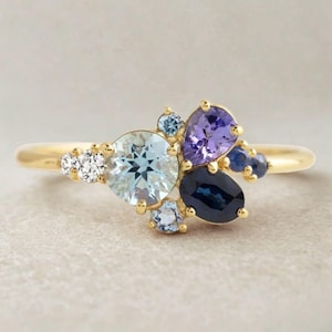 Pode incluir: Um anel de ouro com um conjunto de pedras preciosas. O anel apresenta um esquema de cores azul claro, roxo e azul escuro, com algumas pedras brancas pequenas. As pedras preciosas são colocadas numa banda de ouro.