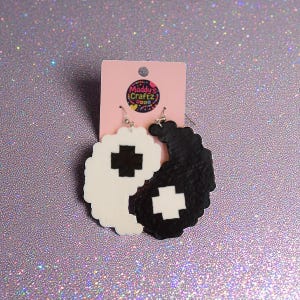 May include: Yin and Yang earrings in black and white, with a cross design in the center of each earring. The earrings are attached to a pink tag with the text "Maddy's Craftz".