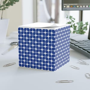 Puede incluir: Un bloc de notas cúbico con un patrón floral azul y blanco. El bloc de notas está sobre un escritorio blanco con clips y un ordenador en el fondo. La parte superior del bloc de notas tiene una pila de papel blanco.