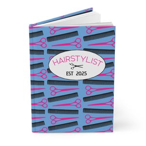 Puede incluir: Un cuaderno azul con un patrón de tijeras rosas y peines negros. La portada presenta el texto "HAIRSTYLIST" en un óvalo blanco, con "EST 2025" debajo. El cuaderno está abierto, revelando páginas blancas en blanco.