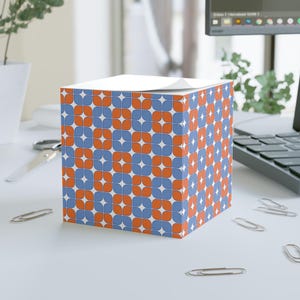May include: A cube-shaped notepad with a repeating pattern of orange and blue floral shapes on a white background. The notepad is sitting on a white desk with paper clips scattered around.
