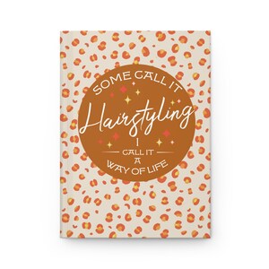 Puede incluir: Un diario de tapa dura con un fondo crema y un patrón repetido de manchas de leopardo naranja. El centro presenta un círculo marrón con las palabras "SOME CALL IT Hairstyling I CALL IT A WAY OF LIFE."
