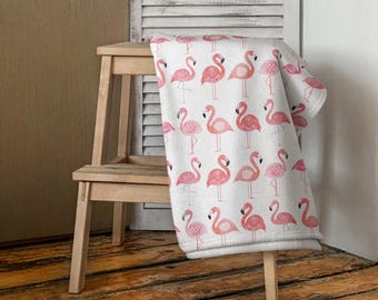 Op de afbeelding: Een witte deken met een roze flamingo-patroon. De deken is gevouwen en over een houten opstapje gelegd.