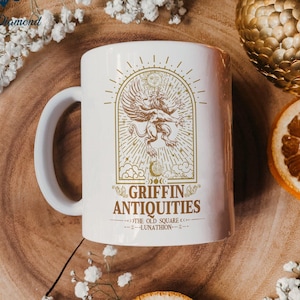 May include: White ceramic mug with a gold design of a griffin with wings spread, surrounded by stars and moons. The text "Griffin Antiquities" is below the griffin, with the words "The Old Square" and "Lunathon" in smaller font below that.