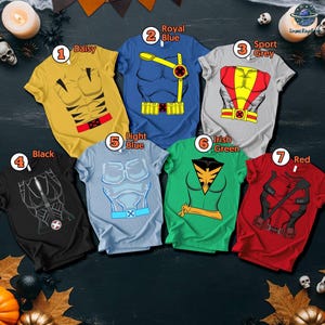 Puede incluir: Siete camisetas de manga corta en varios colores, cada una con un dise&ntilde;o de disfraz de superh&eacute;roe. Los colores incluyen amarillo, azul, gris, negro, azul claro, verde y rojo. Las camisetas est&aacute;n dispuestas sobre una superficie oscura con decoraciones de Halloween.