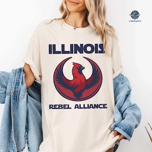 May include: Cream-colored t-shirt with the word "ILLINOIS" in navy blue block letters above a red and navy blue cardinal bird design. The text "REBEL ALLIANCE" is below the bird.
