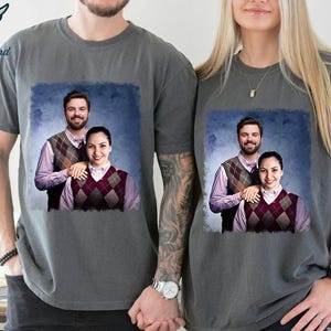 May include: Two grey t-shirts featuring a graphic print of a couple wearing matching jumpers. The couple are standing in front of a blue background.