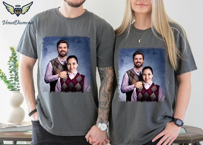 May include: Two grey t-shirts featuring a graphic print of a couple wearing matching jumpers. The couple are standing in front of a blue background.