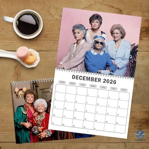 May include: A spiral-bound calendar for December 2026 featuring a photo of four women. The calendar is open to the month of December. A cup of coffee and macarons are on the wooden surface.