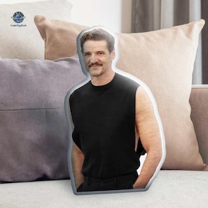 May include: A pillow in the shape of a person wearing a black sleeveless shirt. The pillow is resting on a couch with several other pillows in the background. The person has a mustache and short hair.