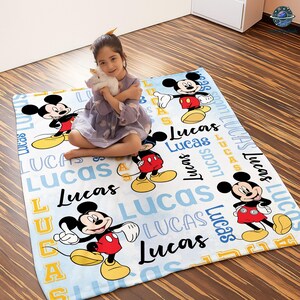A young girl sitting on a Mickey Mouse-themed blanket with the name "Lucas" written on it.