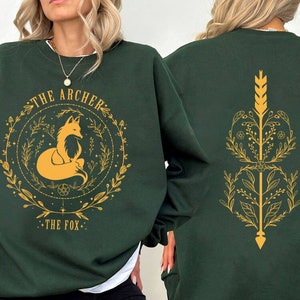 May include: Dark green sweatshirt with a gold fox illustration inside a circular floral design. The text "THE ARCHER" and "THE FOX" are above and below the fox. The back features a gold arrow and floral design.