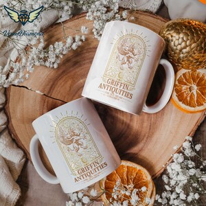 May include: Two white ceramic mugs with a gold design of a griffin and the text "Griffin Antiquities - The Old Square - Lunathrow" on a wooden surface.