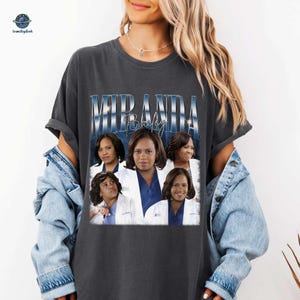May include: Dark gray t-shirt featuring a graphic of Miranda Bailey with the text "MIRANDA Bailey" in a blue and white design. The graphic includes multiple images of the character. The shirt is a casual style.