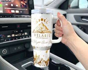 May include: A white insulated tumbler with a handle and straw, featuring gold-coloured illustrations of mountains, an open book, moon phases, and stars. The text reads Velaris City of Starlight and The Night Court City in a gold script font.