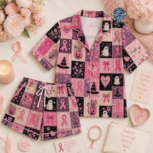 May include: A short-sleeve button-down pajama top and matching shorts set featuring a patchwork pattern of pink and black squares. The design includes motifs of pink awareness ribbons, ghosts, witch hats, pumpkins, and bows with the word hope.