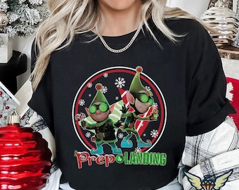 May include: Black t-shirt with a circular graphic of two cartoon elves in green hats and glasses, holding candy canes and gingerbread men. The graphic includes snowflakes and the text "Prep & Landing" in green and red.