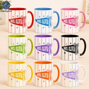May include: Nine ceramic mugs with vertical stripes and a pennant graphic featuring the text MRS. OLIVIA and a checkered bow. Each mug has a colored handle and interior rim, including red, blue, pink, lime green, purple, black, yellow, orange, and white.