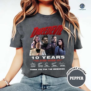 May include: A gray t-shirt featuring the word "DAREDEVIL" in red, with images of characters and the text "10 YEARS 2015-2025". The shirt also includes signatures and the phrase "THANK YOU FOR THE MEMORIES".