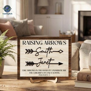 May include: A rectangular wooden sign with a dark brown frame and a light wood grain center. The text reads RAISING ARROWS, followed by the names Smith and Janet with arrow graphics, and the verse LIKE ARROWS IN THE HAND OF A WARRIOR ARE THE CHILDREN OF ONE'S YOUTH. PSALM 127:3-5.