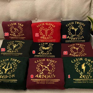 May include: Twelve different t-shirts with gold lettering and graphics. Each shirt has a number and a name of a Greek god or goddess. The shirts are red, black, green, and burgundy.