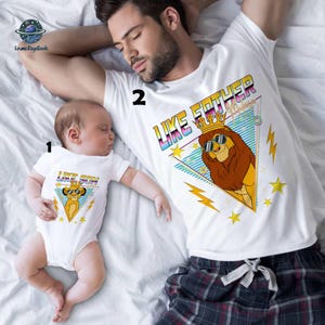 May include: White t-shirts with matching graphics. One shirt says "LIKE FATHER" with a lion wearing sunglasses and a crown. The other shirt says "LIKE SON" with a similar lion graphic. The shirts are on a white background.