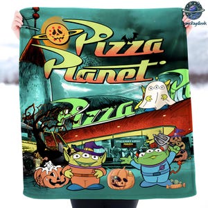 May include: A teal blanket featuring a Pizza Planet design with a spooky Halloween theme. The design includes the Pizza Planet logo, aliens, pumpkins, and a bat against a night sky backdrop. The blanket is held up by a person.