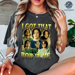 May include: Dark gray t-shirt with the text "I GOT THAT BOB IN ME" in gold lettering. The shirt features a graphic of a man in various poses. The design has a vintage, distressed look.