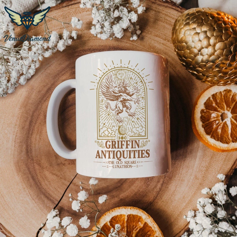May include: White ceramic mug with a gold design of a griffin with wings spread, surrounded by stars and moons. The text "Griffin Antiquities" is below the griffin, with the words "The Old Square" and "Lunathon" in smaller font below that.