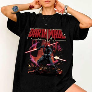 May include: Black t-shirt featuring a graphic of Darth Maul wielding two red lightsabers. The design includes the text "Darth Maul" in red and white, with a background of flames and purple energy.