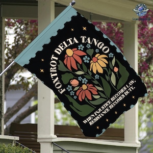 May include: A black garden flag with a blue border. The flag features a stylized floral design with the text "FOXTROT DELTA TANGO" in a circle. The text "WHEN INJUSTICE BECOMES LAW, RESISTANCE BECOMES DUTY." is printed at the bottom of the flag.