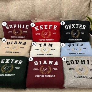 May include: Nine different colored sweatshirts with the text "Foxfire Academy" and a crest with the letter "F" in gold. The sweatshirts are labeled with numbers 1 through 6 and the names Sophie, Keefe, Dexter, Biana, Tam, and Fitz.