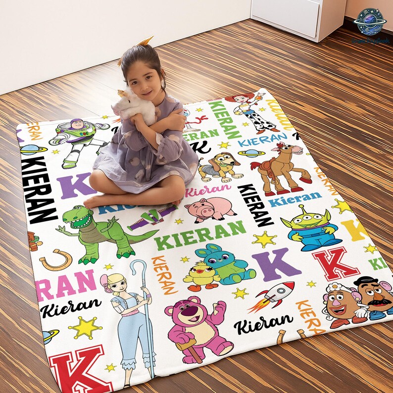 A young girl is sitting on a colorful blanket with various cartoon characters and characters from the Toy Story movie.