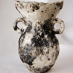 May include: A small, hand-formed ceramic vase with a wide, flared rim and two circular side handles. The surface features a heavily textured, mottled finish with a stark contrast between deep black patches and a weathered, off-white base color.