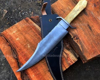 May include: A large, silver-bladed knife with a bone handle rests in a black leather sheath. The knife has a serrated edge and is set against a backdrop of rough-hewn wood. The sheath is stitched with white thread.