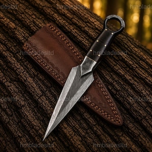 Può includere: Un pugnale artigianale con una lama in acciaio a motivi e un manico in legno scuro con dettagli in ottone. Il pugnale poggia su una superficie di legno strutturata, accompagnato da un fodero in pelle marrone.