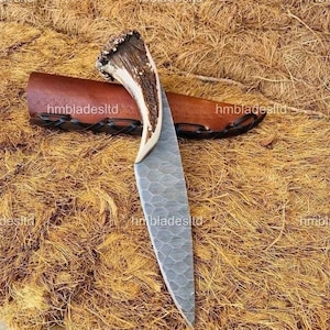 Peut inclure: Un couteau de chasse artisanal avec une lame texturée et un manche en corne de cerf. Il est accompagné d'un étui en cuir marron. La lame présente un motif unique, et le manche a un aspect naturel et rustique. Le texte "hmbladesltd" est visible sur l'étui.