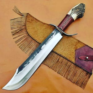 Puede incluir: Un cuchillo grande, artesanal, con una hoja de acero pulido y un mango de asta y cuero rojo oscuro. El cuchillo descansa sobre una funda de cuero marrón con flecos.