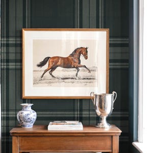 May include: A framed equestrian print of a brown horse in a gold frame hangs above a wooden table. The table holds a silver trophy, a blue and white vase, and a stack of books. The wall is covered in a dark green plaid wallpaper.