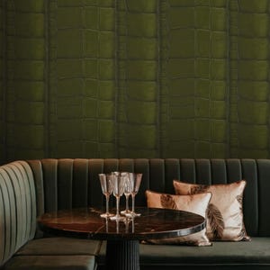 May include: Dark green crocodile-textured wallpaper covers the wall. A dark green velvet booth with a round table and wine glasses is in front of the wall. Decorative pillows with feather designs add a touch of elegance.