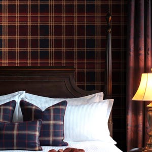 May include: A bedroom scene featuring a dark wooden headboard, white bedding, and plaid accent pillows in navy, red, and tan. The wallpaper and curtains have a matching plaid pattern. A lamp with a cream-colored shade sits on a side table.