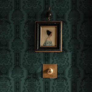 May include: A framed portrait of a person in profile, featuring a dark wooden frame with gold inner trim, hanging from a small black hook. Below the frame is a square brass plate with a central circular knob and a keyhole in the middle.