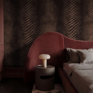 May include: A bedroom scene featuring a bed with a red headboard and gray bedding. A small, round, gray side table holds a mushroom-shaped lamp. The wall behind the bed is covered in a textured, dark brown pattern, and the floor is dark wood.