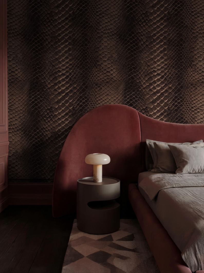 May include: A bedroom scene featuring a bed with a red headboard and gray bedding. A small, round, gray side table holds a mushroom-shaped lamp. The wall behind the bed is covered in a textured, dark brown pattern, and the floor is dark wood.