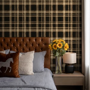 May include: A bedroom scene featuring a brown leather tufted headboard, striped bedding, and decorative pillows. A vase of sunflowers sits on a nightstand next to a lamp. The wall is covered in a black and tan plaid wallpaper.